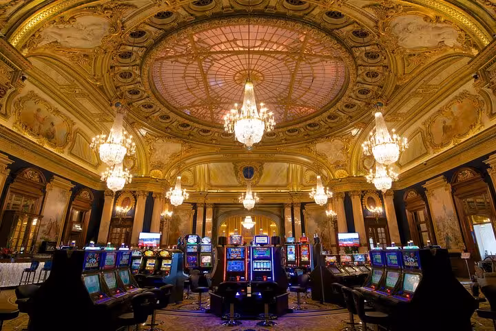 Majestic chandelier-lit gaming hall in Monte-Carlo, highlighting ornate ceiling and slot machines for a lavish night tour.