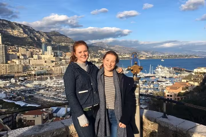 Two tourists enjoy a scenic view of Monte-Carlo's marina and cityscape under a clear blue sky.