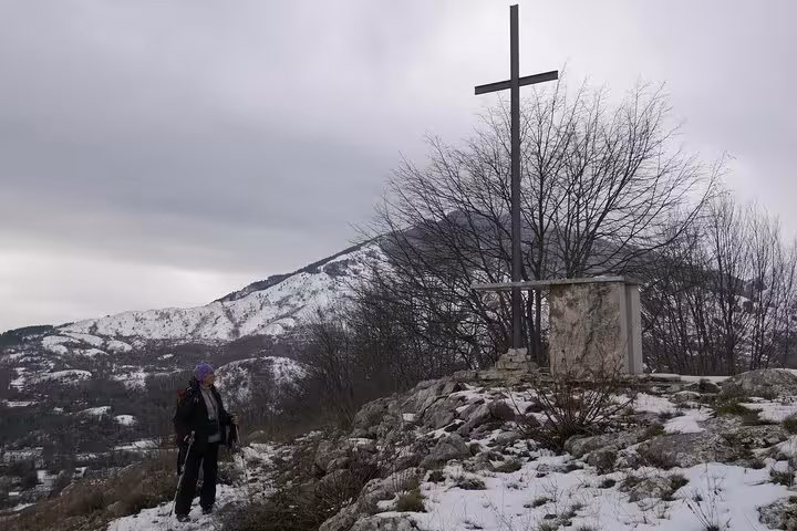 Solo hiker standing beside a war memorial cross on snowy Monte Cassino hilltop with scenic mountain backdrop