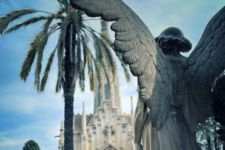 A stone angel statue with outstretched wings stands before palm trees and a historic building in Montjuïc Cemetery.