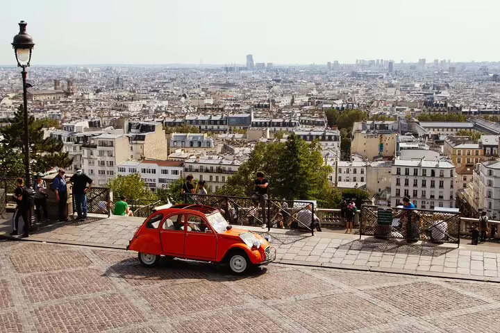 Vintage red Citroën 2CV parked on cobblestone street in Montmartre with Paris cityscape in background