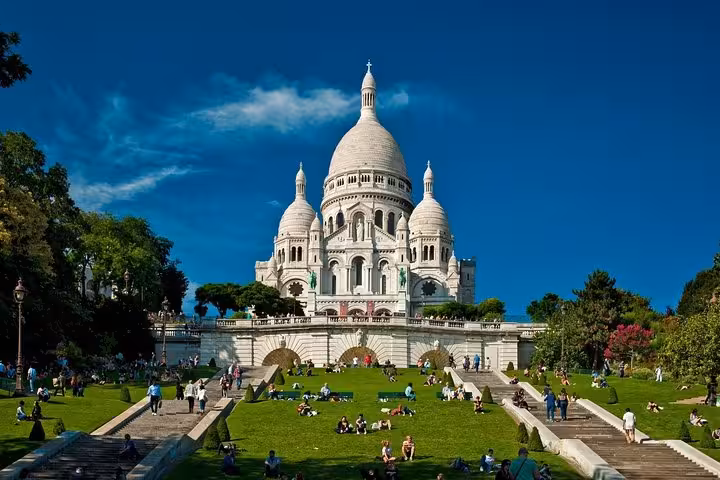 Visitors relax on the lush lawns of the Montmartre hill with the iconic Sacré-Cœur Basilica in the background.