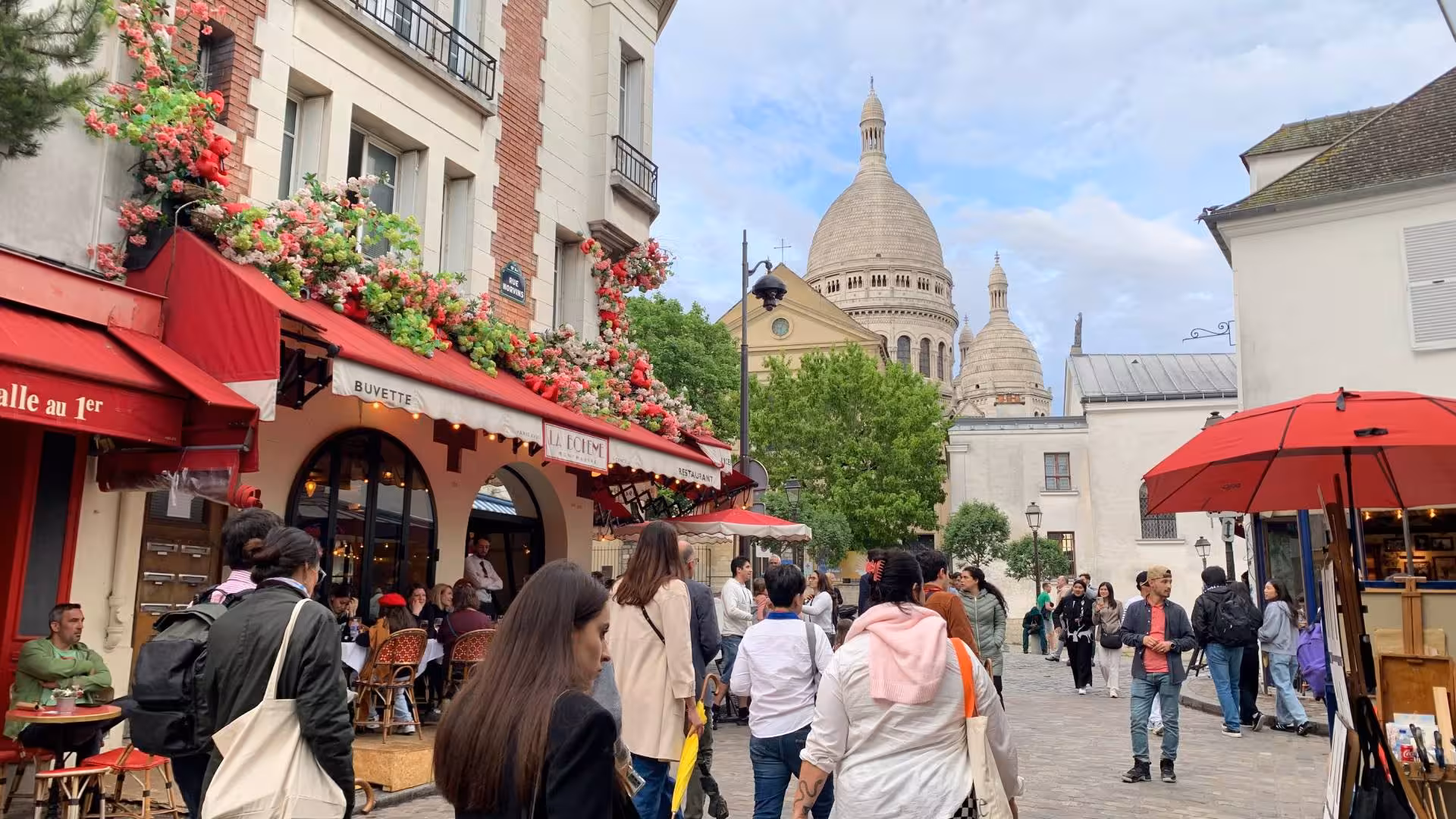 Busy Montmartre square with La Bohème café and Sacré-Cœur basilica dome rising above Parisian streets