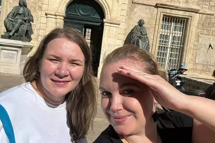 Two women smiling in front of historic statues at a sunlit square in Montpellier, France