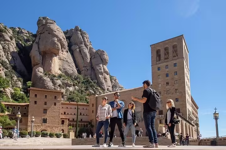 A group of tourists enjoy a sunny day exploring Montserrat Abbey with towering rock formations in the background.