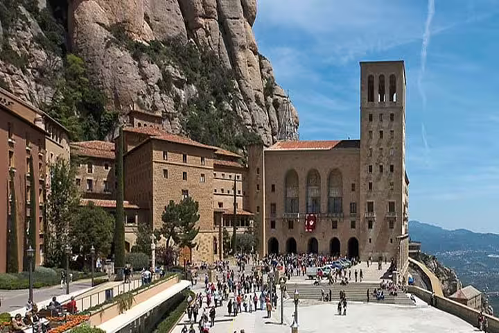 Visitors exploring the historic Montserrat Monastery nestled against rugged mountain cliffs on a sunny day.