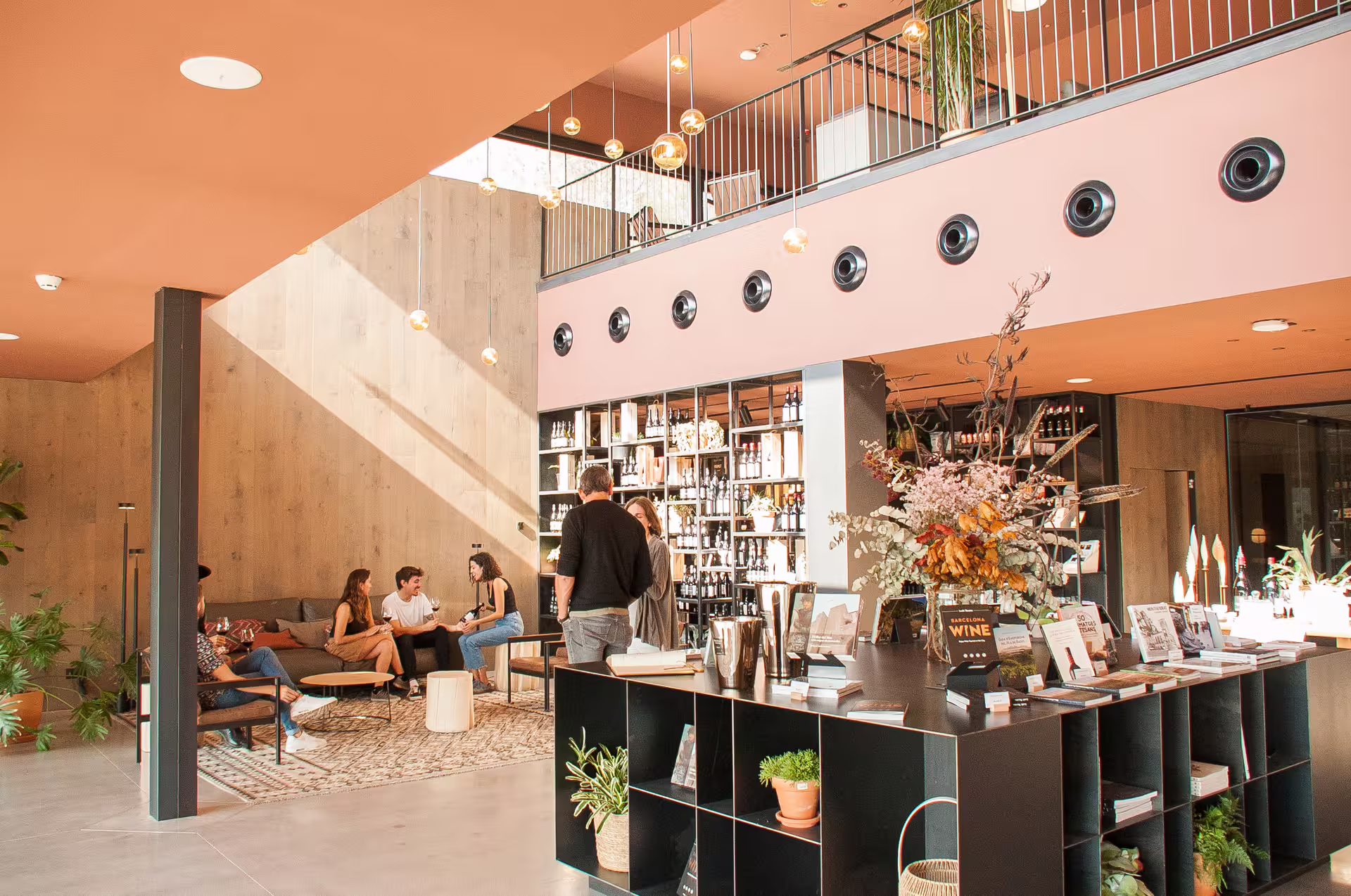 Modern lounge interior with visitors enjoying conversation, surrounded by wine displays, books, and natural decor elements.