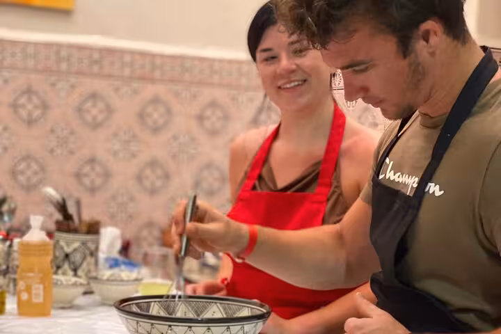 Man whisking ingredients in a bowl while a woman in a red apron smiles during a Moroccan cooking class.