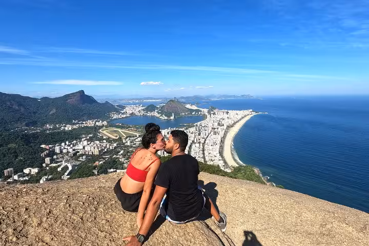 A couple enjoys a kiss atop Two Brothers peak, with panoramic views of Rio's coastline and vibrant cityscape below.