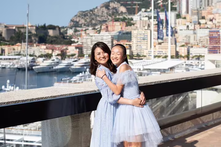 Mother and daughter in elegant dresses embrace with a stunning view of Nice's marina and cityscape in the background.