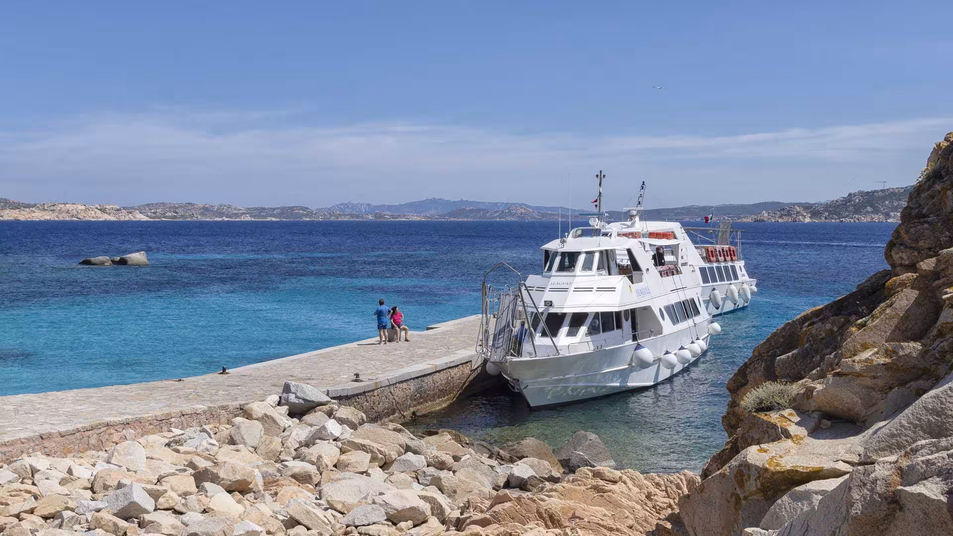 Motorboat moored at rocky pier, offering stunning views of La Maddalena Archipelago's azure waters and coastline.