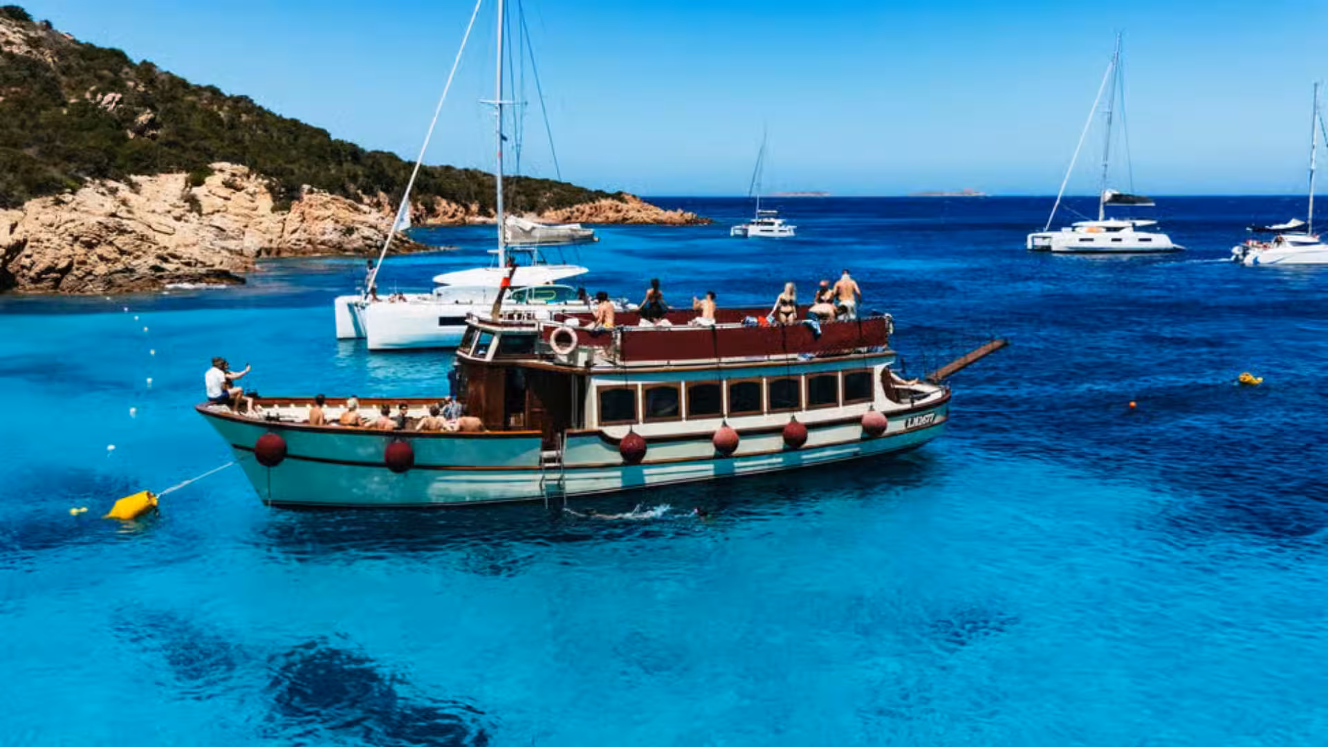 A motorboat anchored in the stunning blue waters of La Maddalena Archipelago, with scenic coastal views and other boats nearby.
