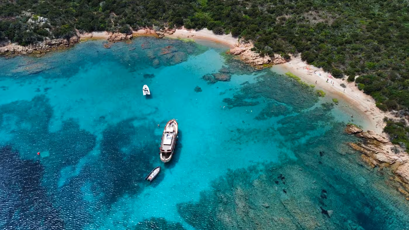 Aerial view of motorboats anchored in the turquoise waters of La Maddalena Archipelago near a sandy beach.