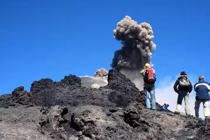 Tourists observe an eruption at Mount Etna, surrounded by rugged volcanic terrain under a clear blue sky.