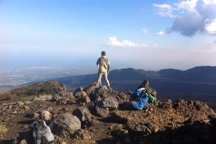 Person standing on rocky summit of Mount Etna, enjoying panoramic views and clear blue skies.
