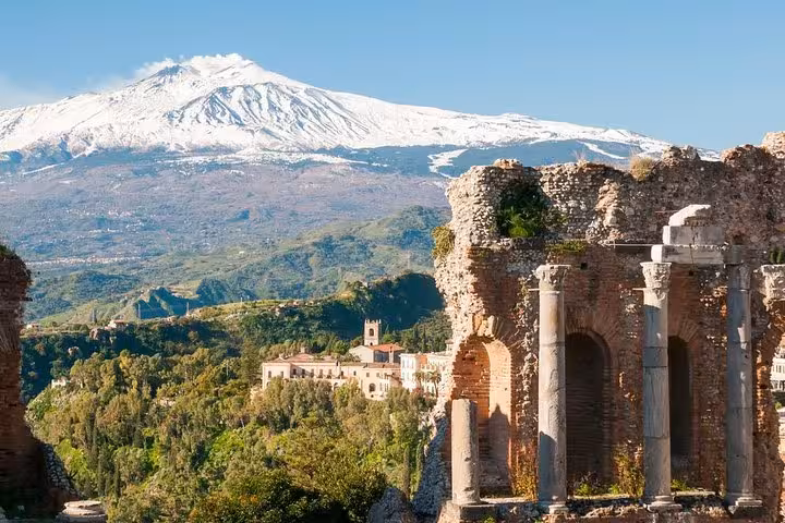Scenic view of Mount Etna behind the ancient ruins of Taormina's Greek theatre amidst lush greenery.