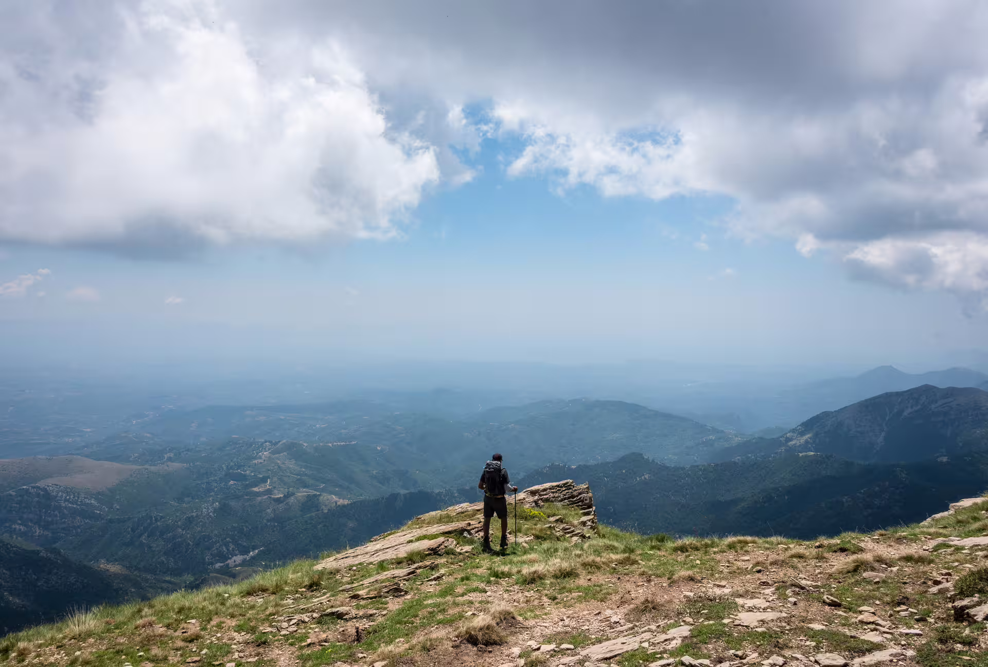 Hiker on Mount Taygetos summit trail at 2407m, overlooking sweeping Peloponnese valleys under dramatic clouds
