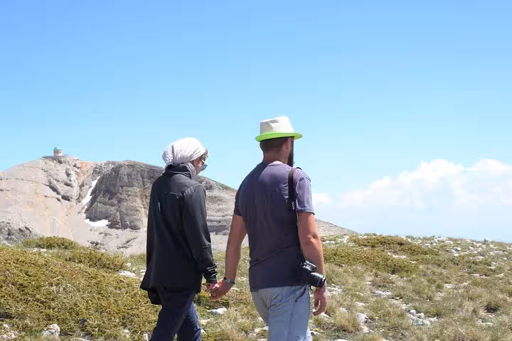 Two hikers walk hand in hand on Mount Tomorr's summit, enjoying clear blue skies and stunning alpine scenery.