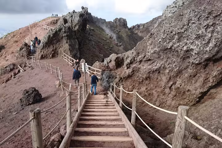 Hikers on Mount Vesuvius crater trail during Naples group tour, climbing steps through volcanic rocks and steam