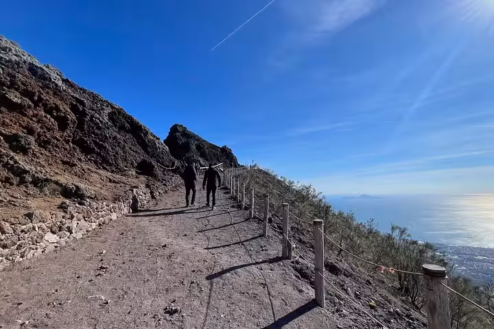 Two hikers ascending the scenic path on Mount Vesuvius with stunning views of the sea and clear blue skies.