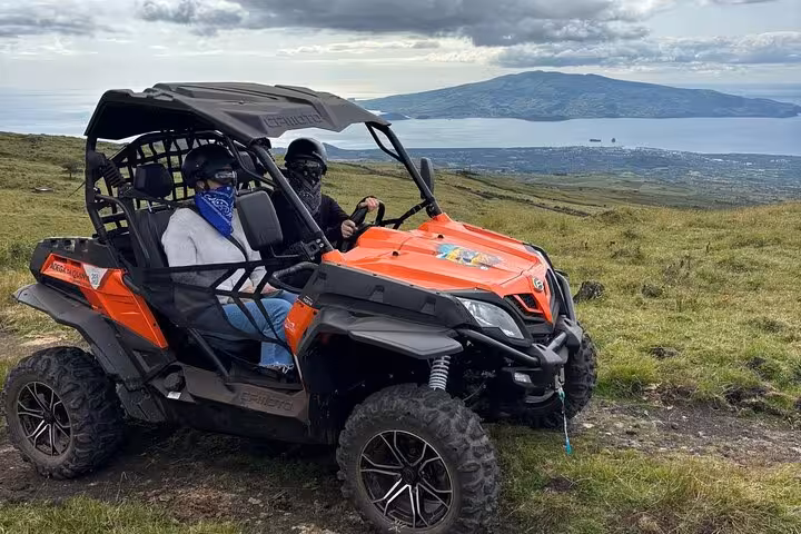 Two riders in an orange buggy explore lush mountain landscapes with panoramic ocean views during a guided tour.