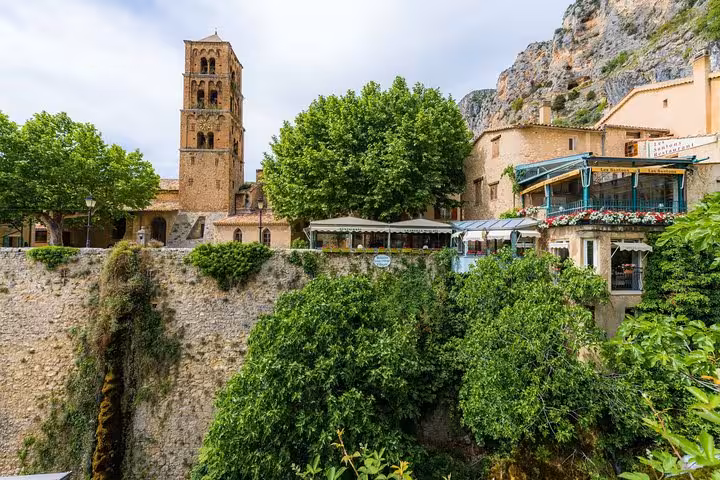 Medieval bell tower and stone buildings nestled among lush green trees beside a rocky cliff in Moustiers-Sainte-Marie