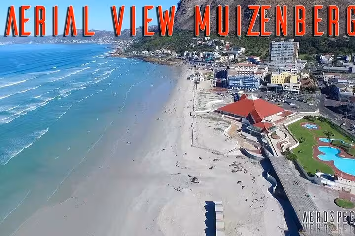 Aerial view of Muizenberg's scenic coastline with waves lapping the shore, vibrant buildings, and mountain scenery.