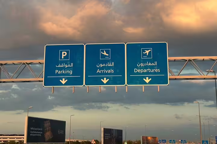 Blue directional signs for parking, arrivals, and departures at Muscat Airport under a dramatic sunset sky.