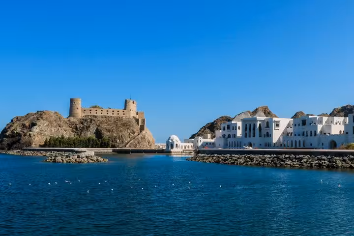 Al Jalali Fort on rocky hill overlooking blue harbor with white buildings in Muscat, Oman