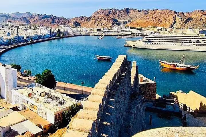 Panoramic view of Muscat's harbor with traditional dhows, luxury yachts, and scenic mountains on a half-day city tour.
