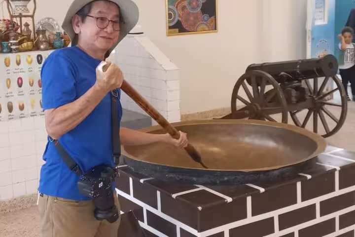 Traveler at Omani heritage stop on private Muscat to Nizwa transfer, posing with giant traditional cooking pan display
