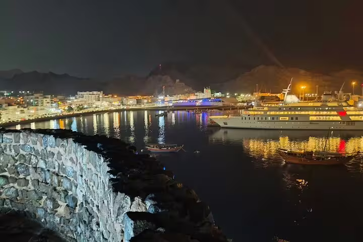 Nighttime view of Muscat harbor featuring docked yachts and city lights reflecting on the water during a private tour.
