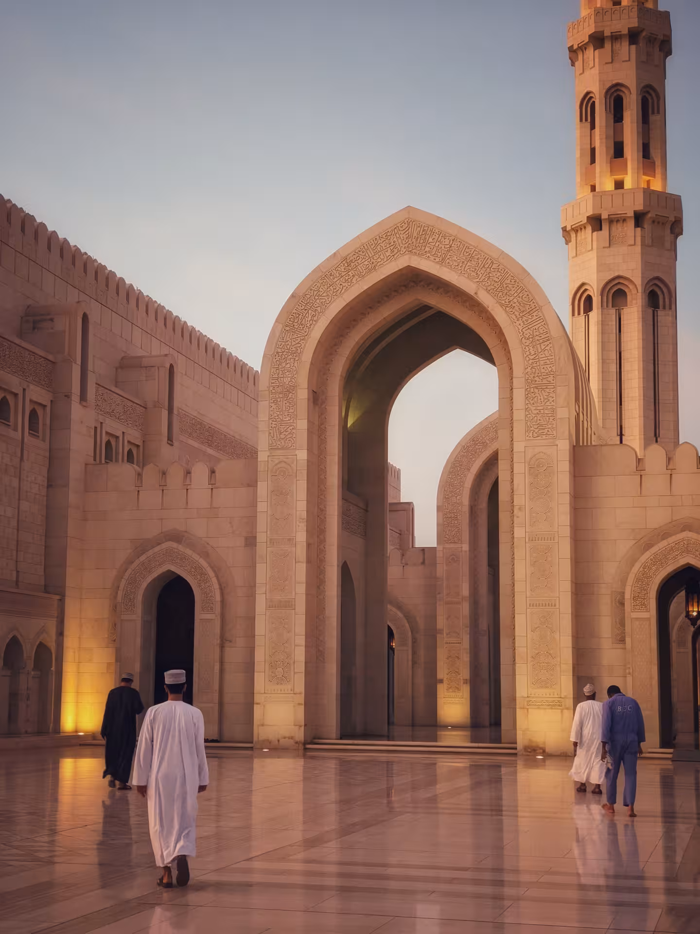 Travelers walk through the illuminated archways of Sultan Qaboos Grand Mosque in Muscat at sunset on an Oman desert tour
