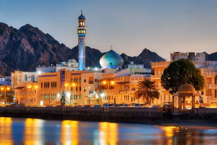 Muscat waterfront at dusk with illuminated mosque minaret, turquoise dome, and mountain backdrop