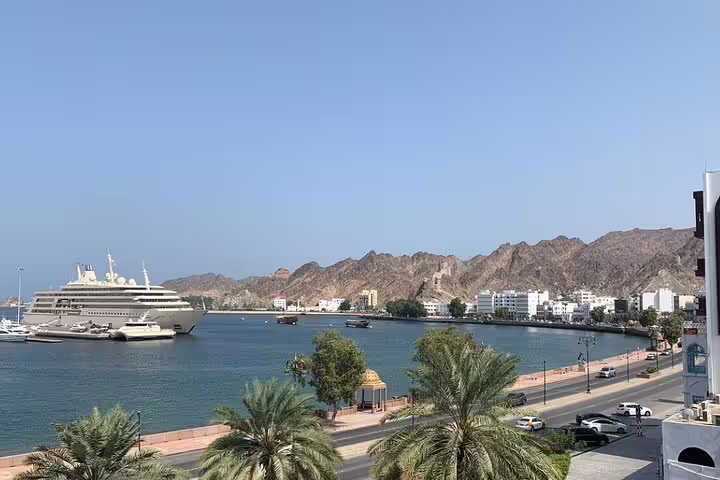 Scenic view of Muscat's waterfront featuring a cruise ship, mountains, and palm-lined promenade on a sunny day.
