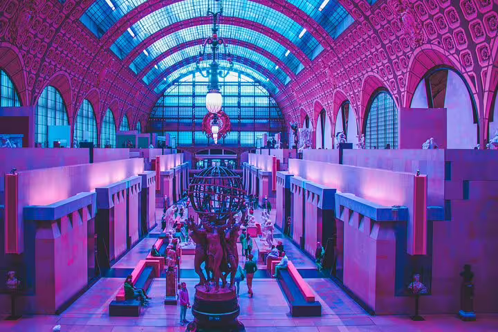 Grand hall of Musée d'Orsay with sculptures, arched glass ceiling, and visitors bathed in purple light