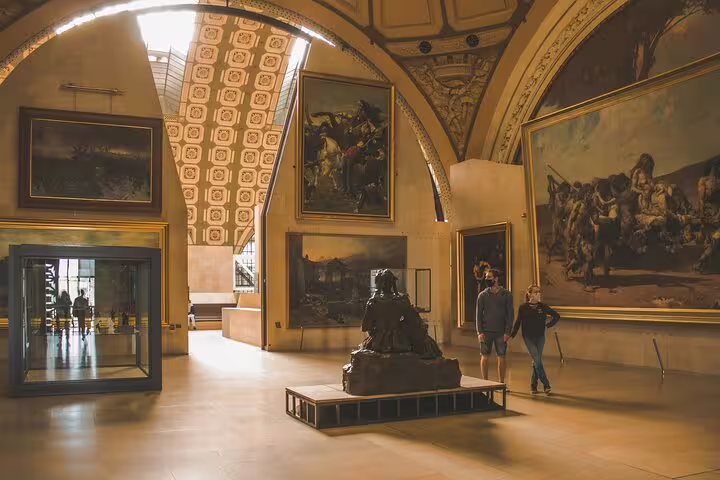 Grand gallery hall at Musée d'Orsay with large paintings, arched ceilings, bronze sculpture, and two visitors