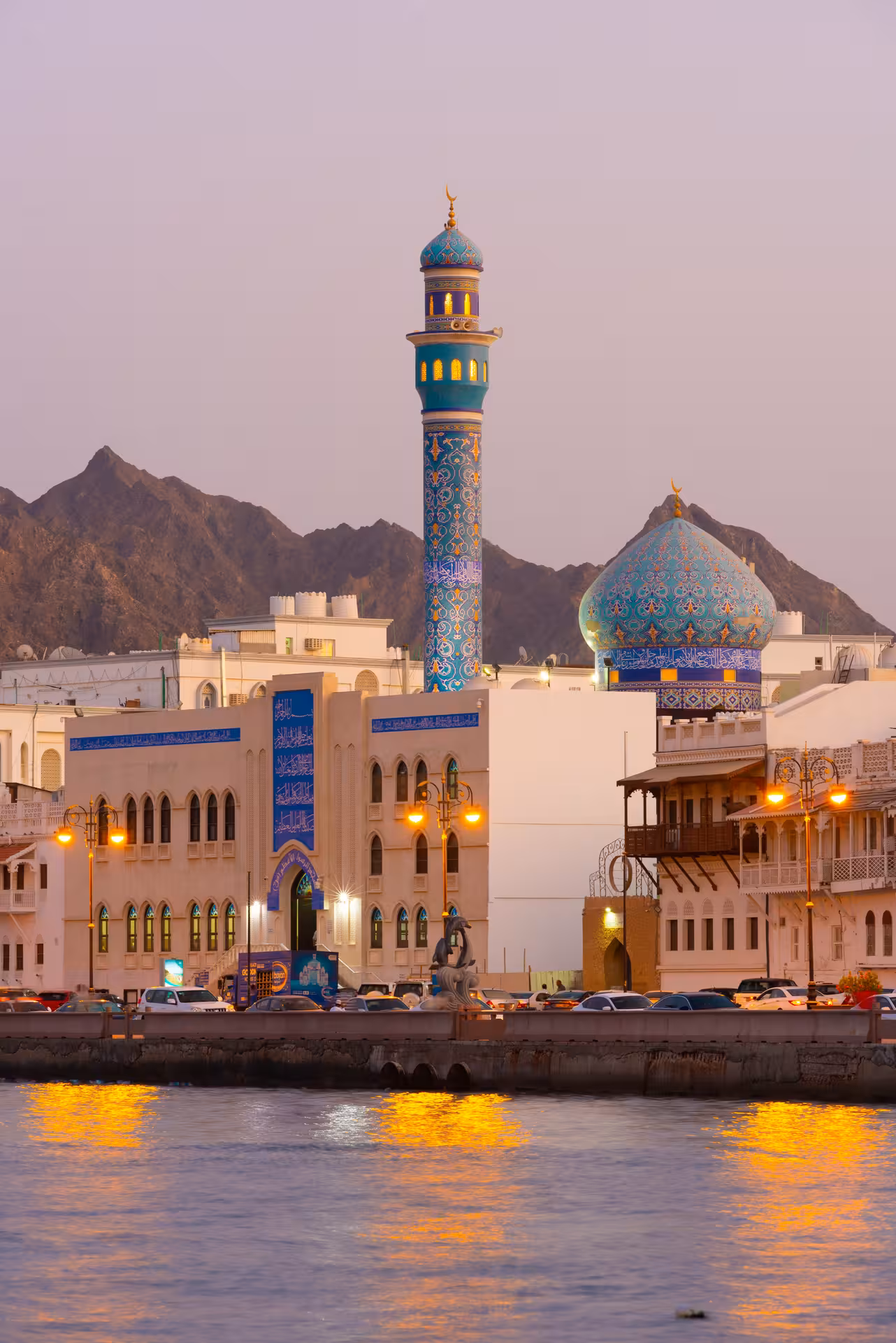 Twilight view of Muttrah Corniche mosque and harbor, with blue minaret, domed roof and mountains on Muscat city tour