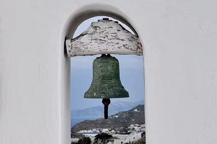 Close-up church bell framed by white arch with Mykonos hills and sea, on a private guided island tour