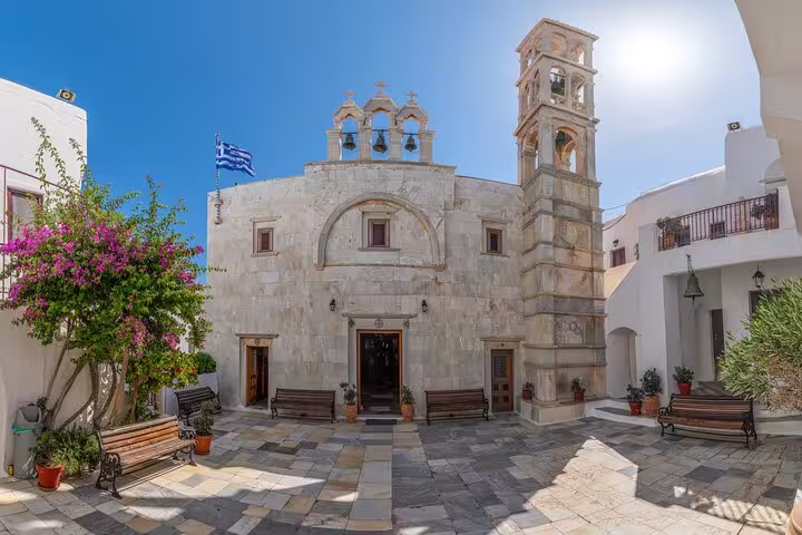 Historic stone church in Mykonos countryside with Greek flag, vibrant flowers, and sunlit courtyard for tasting experience tour.