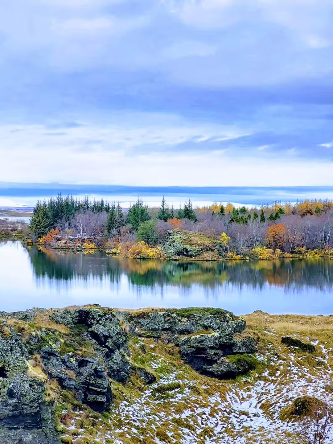 Calm lake reflecting autumn trees and mossy lava rocks at Myvatn, Iceland under a cloudy blue sky