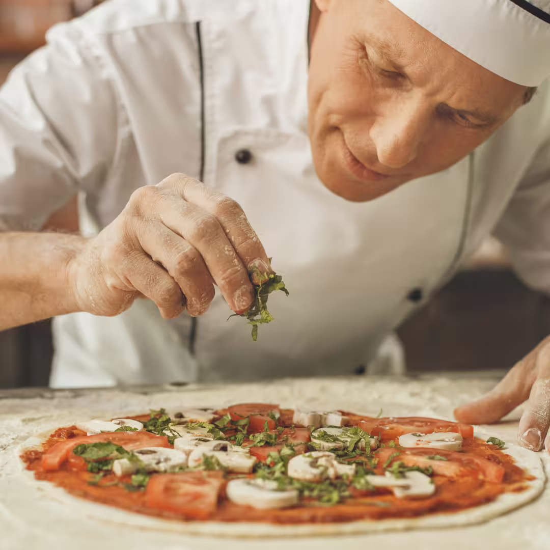 Naples pizza master class chef sprinkling fresh basil on uncooked Neapolitan pizza topped with tomato and mushrooms