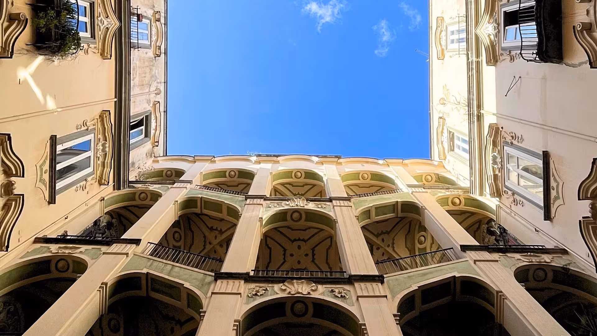 Looking up at the ornate arches of a historic courtyard against a clear blue sky in Naples' Sanità district.