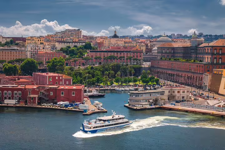 View of Naples waterfront with a boat cruising the harbor, surrounded by historic architecture and lush greenery under a vibrant sky.