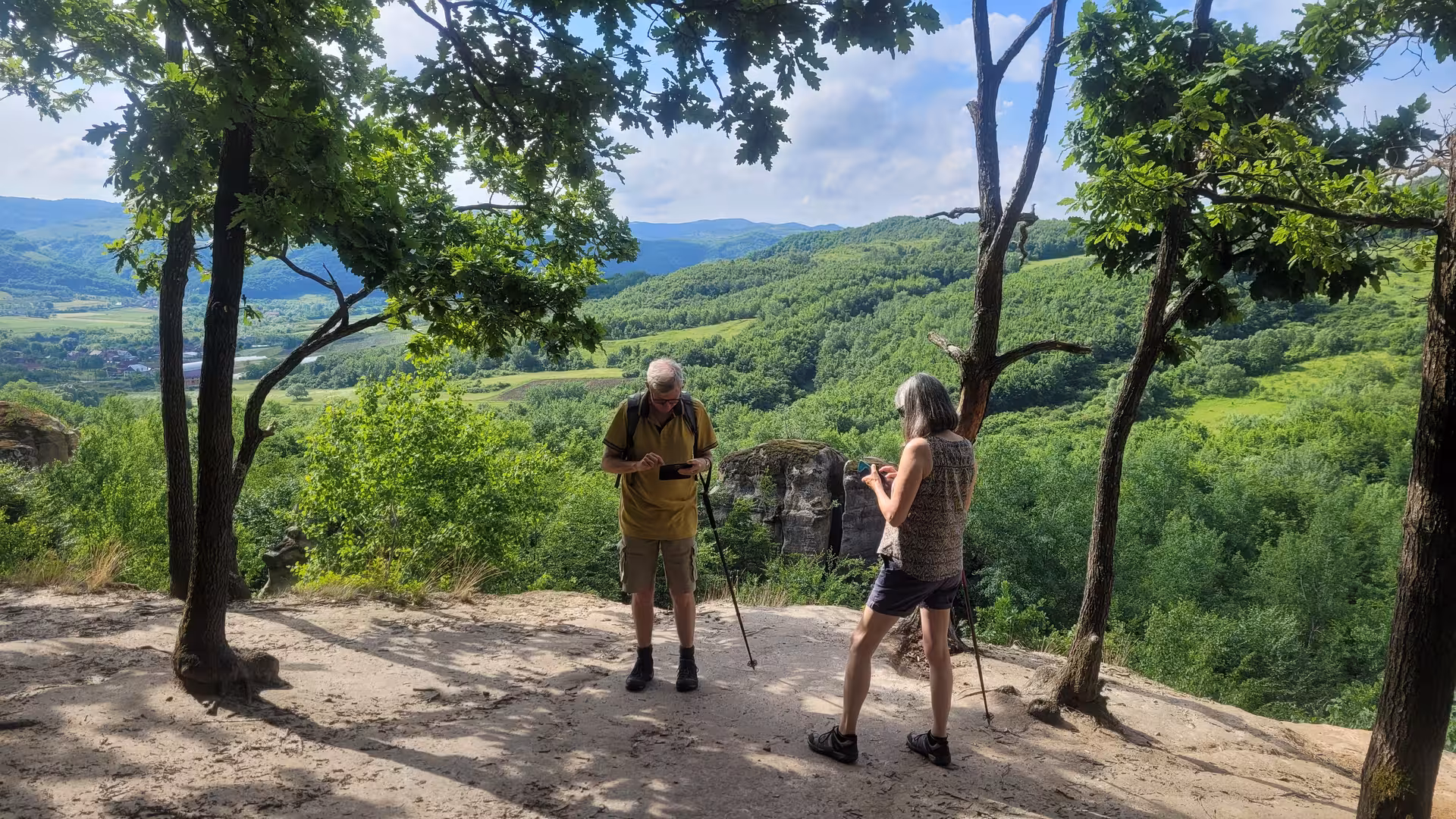 Two hikers with trekking poles on a rocky overlook surrounded by lush green hills in Salaj, Romania
