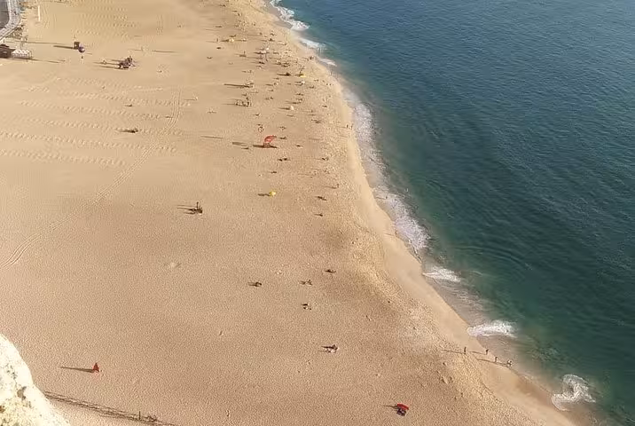 Aerial view of Nazaré's expansive sandy beach with gentle waves meeting the shoreline under a clear blue sky.
