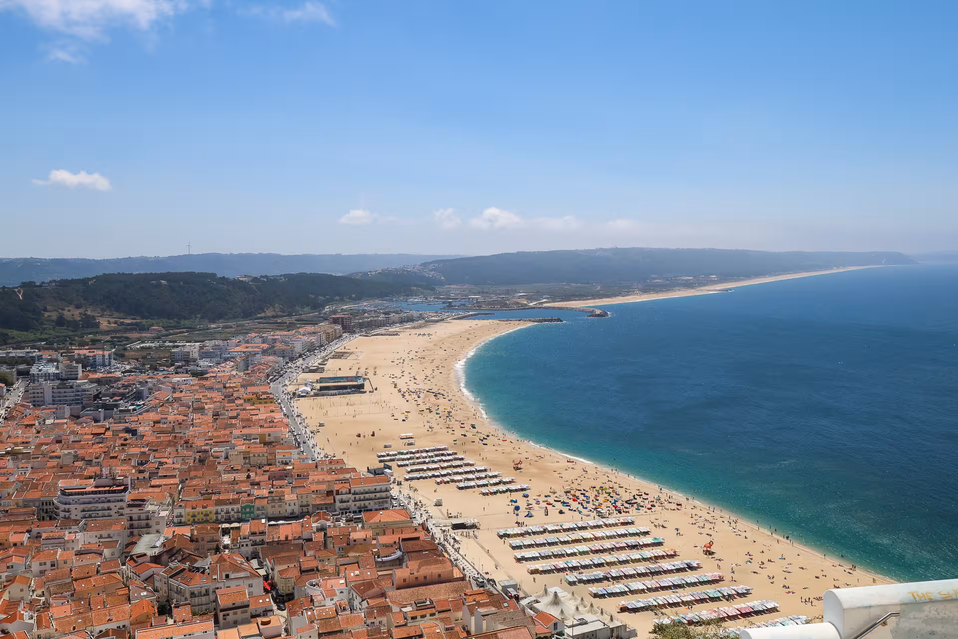 Aerial view of Nazaré's expansive beach and bustling coastline with vibrant umbrellas and azure waters in Portugal.