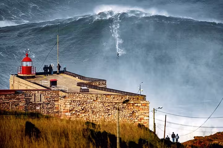 Dramatic view of a surfer riding massive waves near the iconic lighthouse in Nazaré, Portugal, on a Fátima, Nazaré, Batalha & Óbidos tour.