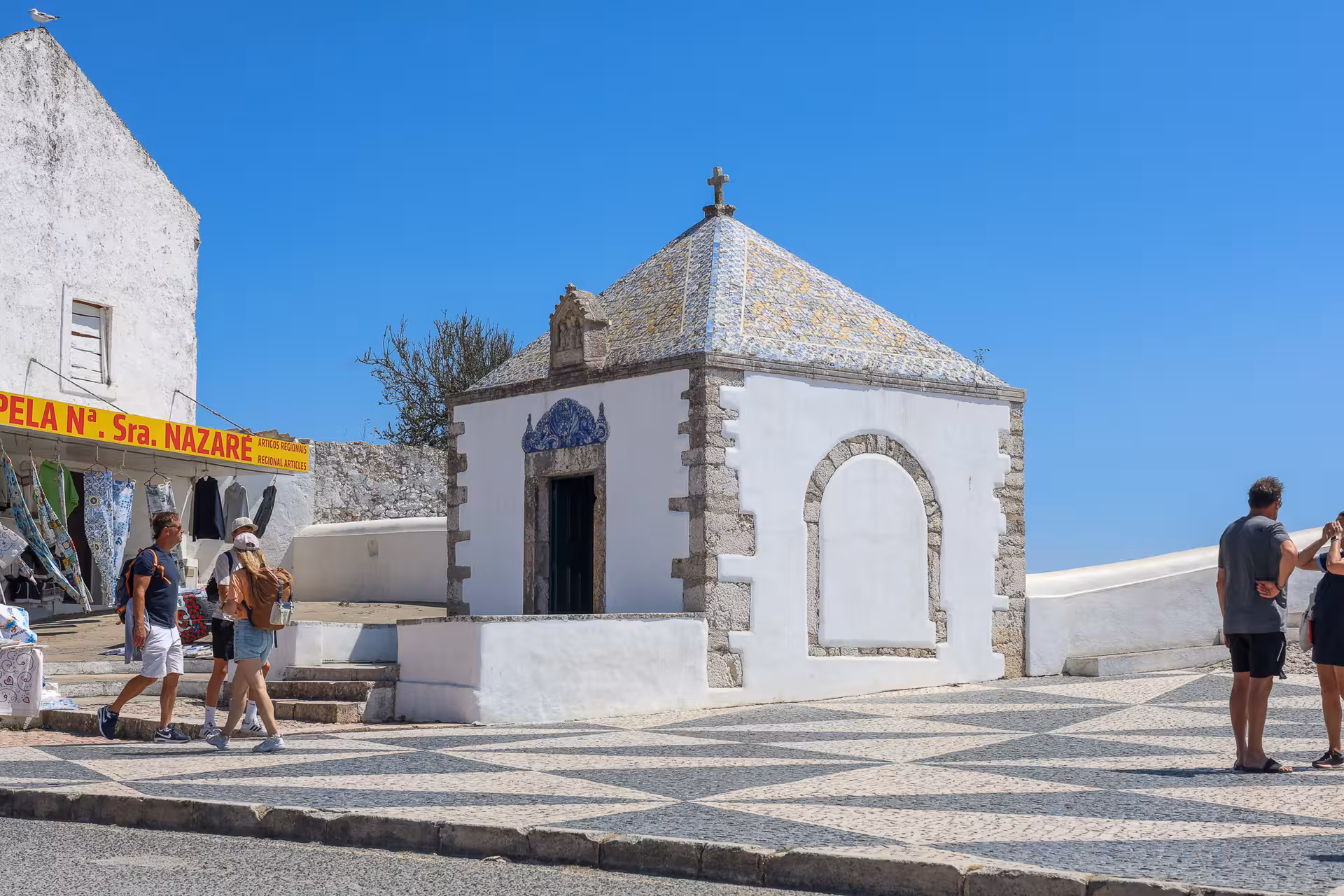 Visitors explore the quaint Chapel of Nossa Senhora in Nazaré, surrounded by traditional shops and vibrant architecture.