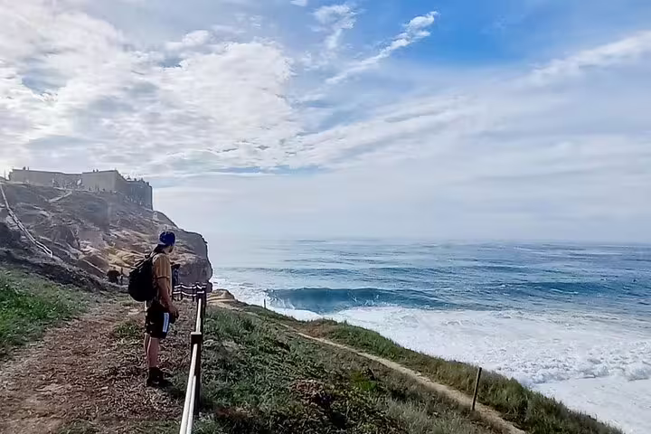 A hiker gazes at the expansive ocean waves from a cliffside path in Nazare, under a picturesque sky.